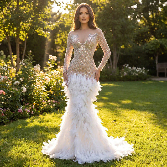 A woman stands in a green garden, wearing a sheer, long-sleeve evening gown[cite: 9] adorned with sparkly silver rhinestones[cite: 9] on the bodice and a dramatic white feather mermaid skirt
