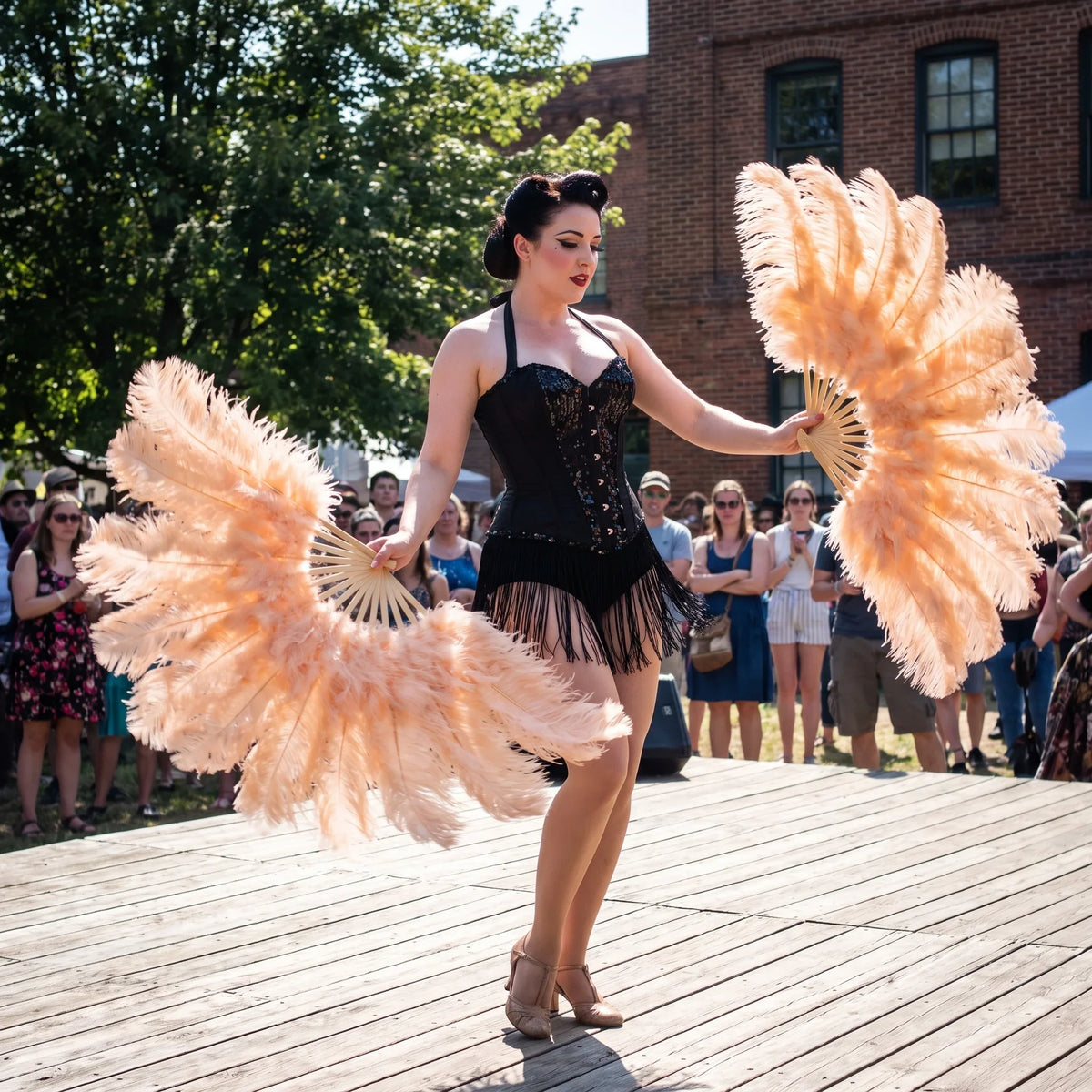 Burlesque dancer in a black corset performing with peach ostrich feather fans on a wooden stage.
