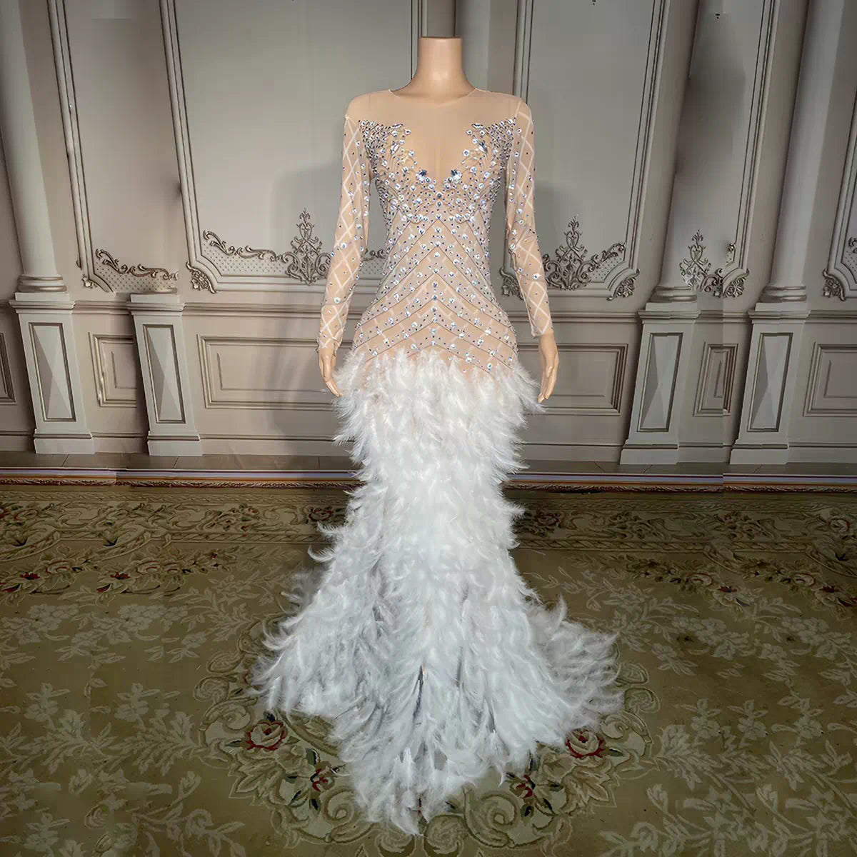A formal mermaid gown displayed on a mannequin in an ornate interior. The dress has a sheer nude-beige bodice with long sleeves and dense silver and blue beading, transitioning into a voluminous white feathered skirt.