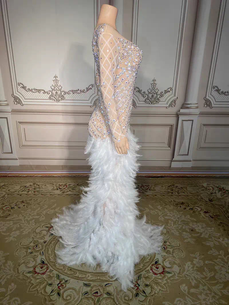 A formal mermaid gown displayed on a mannequin in an ornate room. The dress has a sheer embellished bodice with silver rhinestone-like patterns, long sheer sleeves, and a voluminous white feathered skirt.